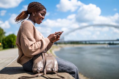 Genç mutlu siyah kadın akıllı telefon kullanıyor. Şehirdeki nehrin kenarında oturuyor..