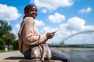 Genç neşeli siyah kadın akıllı telefon kullanıyor. Şehirdeki nehrin kenarında oturuyor..