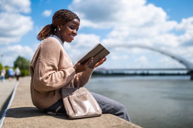 Genç siyah kadın şehirde nehrin kenarında oturuyor. Rahatlıyor ve kitap okuyor..