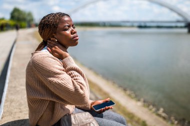 Genç ve güzel siyahi kadın şehirde nehrin kenarında oturuyor. Boynunda ağrı var..