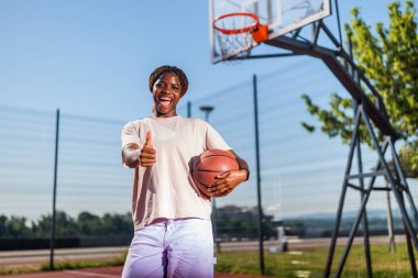 Genç bir kadın sabah spor salonunda tek başına basketbol oynuyor. Güneşli bir günde basketbol oynayan mutlu kız. Başparmağını kaldırıyor..