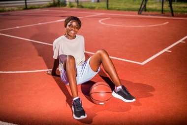 Genç siyahi kadın sabahları spor salonunda tek başına basketbol oynuyor. Mutlu kız güneşli bir günde basketbol sahasında oturuyor..
