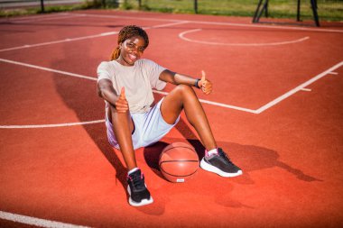Genç siyahi kadın sabahları spor salonunda tek başına basketbol oynuyor. Mutlu kız güneşli bir günde basketbol sahasında oturuyor..