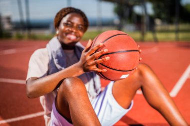 Genç siyah kadın basketbol sahasında oturuyor. O, topu tutuyor. Seçici odak.
