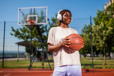 Genç bir kadın sabah spor salonunda tek başına basketbol oynuyor. Mutlu kız basketbol oynuyor ve güneşli bir günde müzik dinliyor..