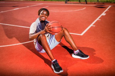 Genç siyahi kadın sabahları spor salonunda tek başına basketbol oynuyor. Mutlu kız güneşli bir günde basketbol sahasında oturuyor..