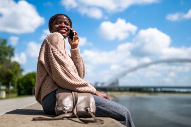 Genç, neşeli siyahi kadın telefonda konuşuyor. Gülüyor ve şehirde güneşli bir günün tadını çıkarıyor..