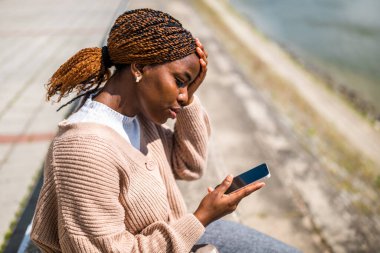 Genç endişeli siyah kadın akıllı telefon kullanıyor. Şehirdeki nehrin kenarında oturuyor..