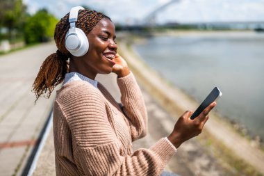 Genç siyah kadın akıllı telefon kullanıyor ve kulaklıkla müzik dinliyor, nehir kenarında..