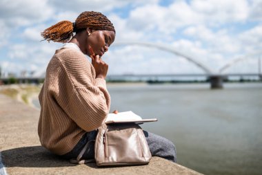 Genç siyah kadın şehirde nehrin kenarında oturuyor. Rahatlıyor ve planlar yapıyor..