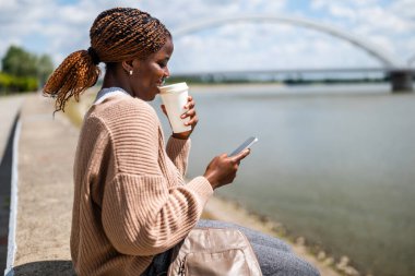 Genç siyah kadın nehir kenarına gitmek için akıllı telefon kullanıyor ve kahve içiyor..