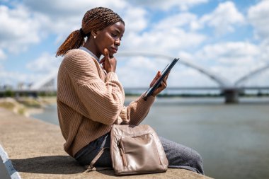 Genç endişeli siyah kadın dijital tablet kullanıyor. Şehirdeki nehrin kenarında oturuyor..