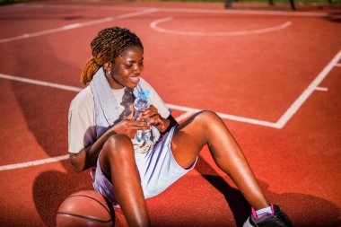 Genç siyahi kadın sabahları spor salonunda tek başına basketbol oynuyor. Elinde bir şişe su tutuyor ve mola veriyor..