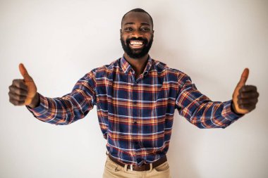 Rahat gömlekli mutlu siyah adam duvarın önünde duruyor ve gülümsüyor. Başparmağını kaldırıyor..