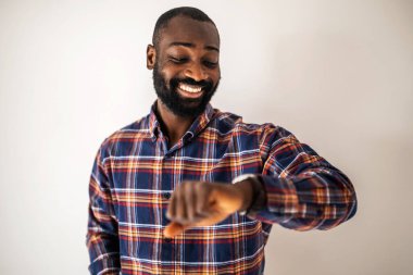 Rahat gömlekli neşeli siyah adam duvarın önünde duruyor ve kol saatine bakıyor..