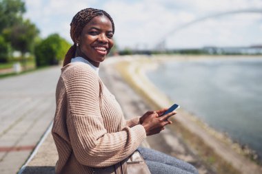 Genç neşeli siyah kadın akıllı telefon kullanıyor. Şehirdeki nehrin kenarında oturuyor..