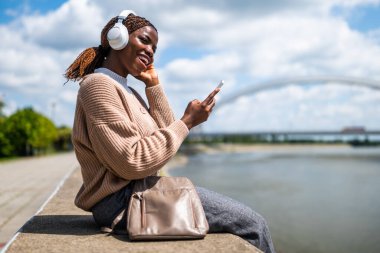 Genç siyah kadın akıllı telefon kullanıyor ve kulaklıkla müzik dinliyor, nehir kenarında..