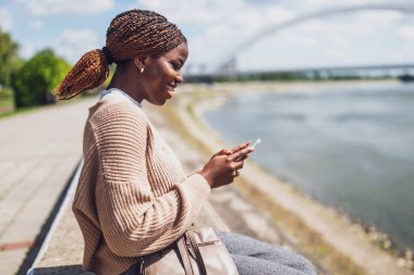 Genç mutlu siyah kadın akıllı telefon kullanıyor. Şehirdeki nehrin kenarında oturuyor..