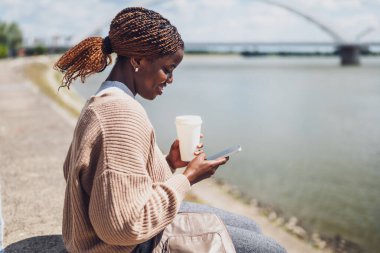 Genç siyah kadın nehir kenarına gitmek için akıllı telefon kullanıyor ve kahve içiyor..