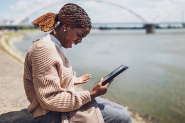 Genç stresli siyah kadın dijital tablet kullanıyor. Şehirdeki nehrin kenarında oturuyor..