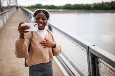 Mutlu genç siyah kadın telefonuna bakıyor ve gülümsüyor. Açık havada güneşli bir günün tadını çıkarıyor ve akıllı telefon kullanarak müzik dinliyor..