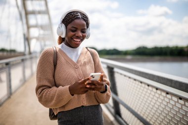 Mutlu genç siyah kadın telefonunda daktilo ediyor ve gülümsüyor. Açık havada güneşli bir günün tadını çıkarıyor ve akıllı telefon kullanarak müzik dinliyor..