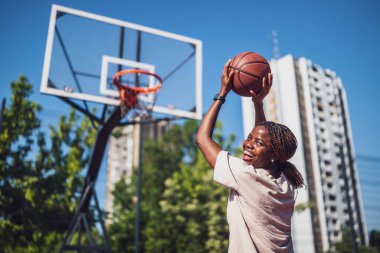 Genç bir kadın sabah spor salonunda tek başına basketbol oynuyor. Kameraya bakıyor ve gülümsüyor..