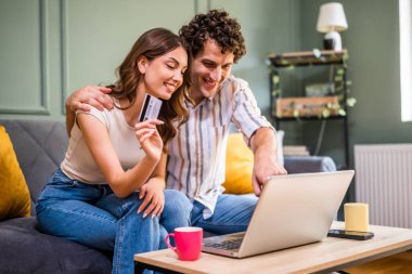 Yetişkin çift evdeki kanepede oturuyor ve dizüstü bilgisayar kullanıyorlar. İnternetten alışveriş yapıyorlar. Kadının elinde kredi kartı var..