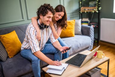 Yetişkin çift evdeki kanepede oturuyor ve dizüstü bilgisayar kullanıyorlar. İnternetten alışveriş yapıyorlar ve alışveriş listesi yapıyorlar..