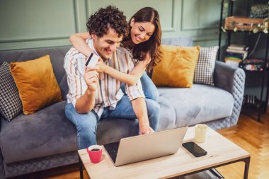 Yetişkin çift evdeki kanepede oturuyor ve dizüstü bilgisayar kullanıyorlar. İnternetten alışveriş yapıyorlar. Adamın elinde ödeme kartı var..