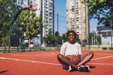 Genç Afrikalı Amerikalı kadın güneşli şehir basketbol sahasında oturuyor, neşeli bir gülümsemeyle bir top tutuyor, neşeyi ve aktif yaşam tarzını temsil ediyor..