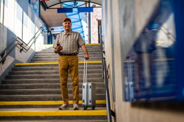 Happy senior man is standing at railway station and messaging on smartphone. Senior people traveling.