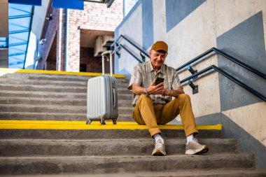 Happy senior man is sitting at railway station and waiting for arrival of the train. He is using digital tablet. Senior people traveling.