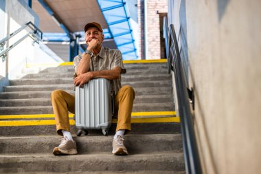 Bored senior man is sitting on bench at railway station and waiting for boarding the train. Senior people traveling.