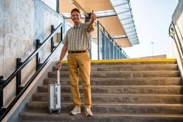 Cheerful senior man is standing at railway station and waiting for arrival of the train. Senior people traveling.