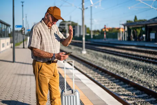 Anxious senior man is standing at railway station and waiting for arrival of the train. Senior people traveling.