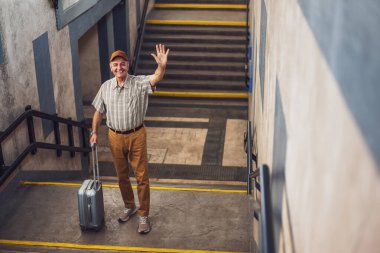 Happy senior man is standing at railway station and waving. He is waiting for arrival of the train. Senior people traveling.