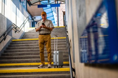 Happy senior man is standing at railway station and messaging on smartphone. Senior people traveling.