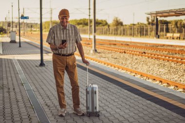 Happy senior man is standing at railway station and messaging on smartphone. Senior people traveling.