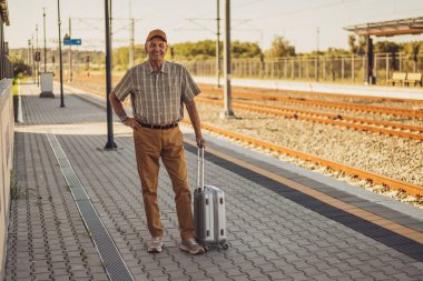 Cheerful senior man is standing at railway station and waiting for arrival of the train. Senior people traveling.
