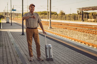 Anxious senior man is standing at railway station and looking at his wristwatch. He is waiting for arrival of the train. Senior people traveling.