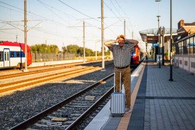Senior man is standing at railway station. He missed the train departure and holding his head in disbelief. Senior people traveling.