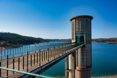 Alentejo Portugal. 04 august 2024.Concrete water tower and walkway over a scenic lake surrounded by hills under a clear blue sky, Santa Clara dam in alentejo Portugal clipart