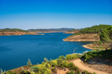 Santa Clara barajının berrak mavi suları, çevreleyen tepeleri ve parlak gökyüzünün altındaki yemyeşil yeşil manzarası, Alentejo Portekiz 'deki Santa Clara barajı.