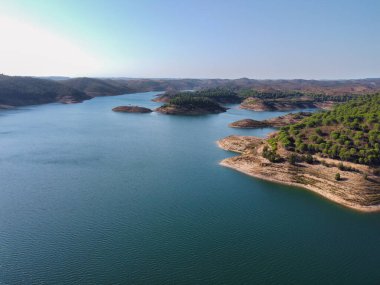 Santa Clara barajının berrak mavi suları, çevreleyen tepeleri ve parlak gökyüzünün altındaki yemyeşil yeşil manzarası, Alentejo Portekiz 'deki Santa Clara barajı.
