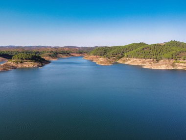 Santa Clara barajının berrak mavi suları, çevreleyen tepeleri ve parlak gökyüzünün altındaki yemyeşil yeşil manzarası, Alentejo Portekiz 'deki Santa Clara barajı.