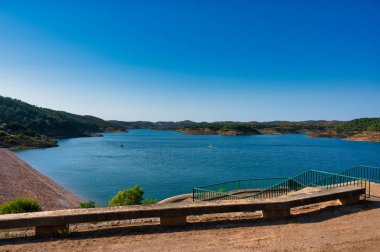 Santa Clara barajının berrak mavi suları, çevreleyen tepeleri ve parlak gökyüzünün altındaki yemyeşil yeşil manzarası, Alentejo Portekiz 'deki Santa Clara barajı.
