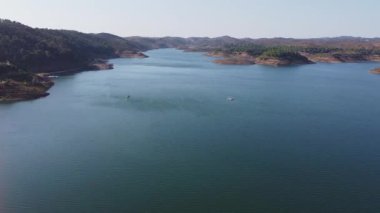 Santa Clara barajının berrak mavi suları, çevreleyen tepeleri ve parlak gökyüzünün altındaki yemyeşil yeşil manzarası, Alentejo Portekiz 'deki Santa Clara barajı.
