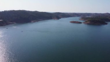 Santa Clara barajının berrak mavi suları, çevreleyen tepeleri ve parlak gökyüzünün altındaki yemyeşil yeşil manzarası, Alentejo Portekiz 'deki Santa Clara barajı.