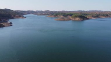 Santa Clara barajının berrak mavi suları, çevreleyen tepeleri ve parlak gökyüzünün altındaki yemyeşil yeşil manzarası, Alentejo Portekiz 'deki Santa Clara barajı.